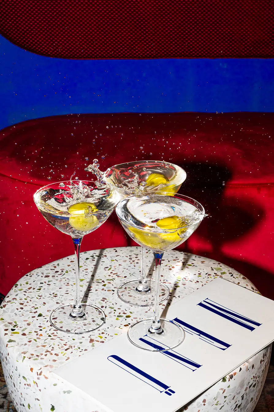 Three martini-style cocktails with yellow drinks sit on a splash plate; liquid and ice splash upward.