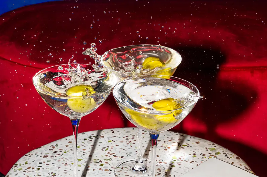 Drei Martini-Style-Cocktails mit gelben Drinks stehen auf einer Spritzschutzfläche; Flüssigkeit und Eis spritzen nach oben.