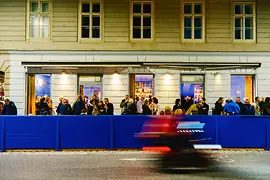 Viele Gäste stehen abends vor der beleuchteten Barfassade; im Vordergrund fährt ein Fahrzeug als rote und blaue Bewegungsunschärfe vorbei.