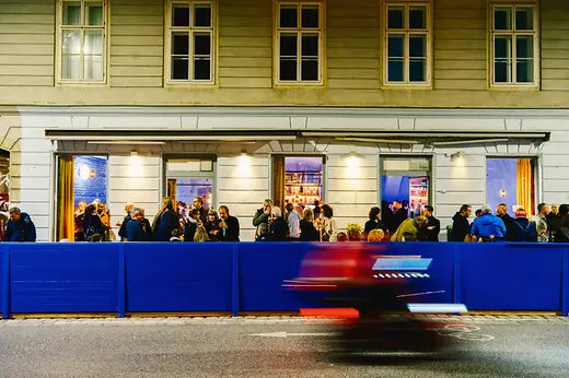 A crowd gathers outside the illuminated bar façade at night, while a passing vehicle creates red and blue motion blur in the foreground.