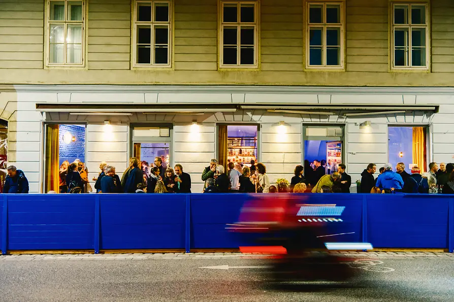Viele Gäste stehen abends vor der beleuchteten Barfassade; im Vordergrund fährt ein Fahrzeug als rote und blaue Bewegungsunschärfe vorbei.