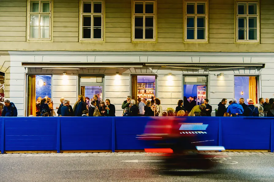 A crowd gathers outside the illuminated bar façade at night, while a passing vehicle creates red and blue motion blur in the foreground.