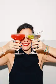 Eine lächelnde Frau hält zwei Cocktails vor ihr Gesicht, sodass die Gläser ihre Augen verdecken.
