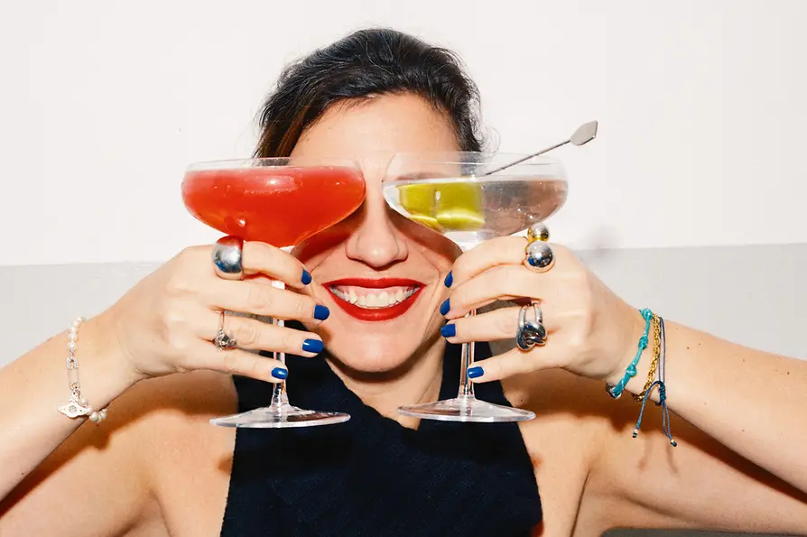Eine lächelnde Frau hält zwei Cocktails vor ihr Gesicht, sodass die Gläser ihre Augen verdecken.