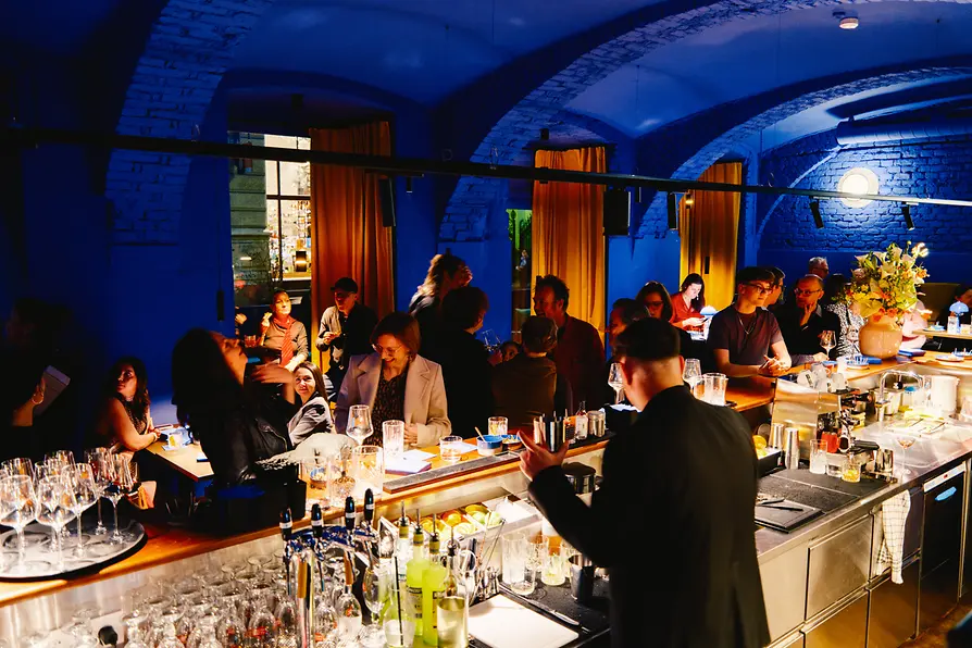 A busy evening dining area with many guests at tables and open doorways between rooms; in the foreground, a bar with bottles and glasses.