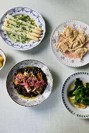 Mehrere Teller mit saisonalen Gerichten stehen auf einem hell gedeckten Tisch, darunter weißer Spargel, gefüllte Pasta und grünes Blattgemüse.