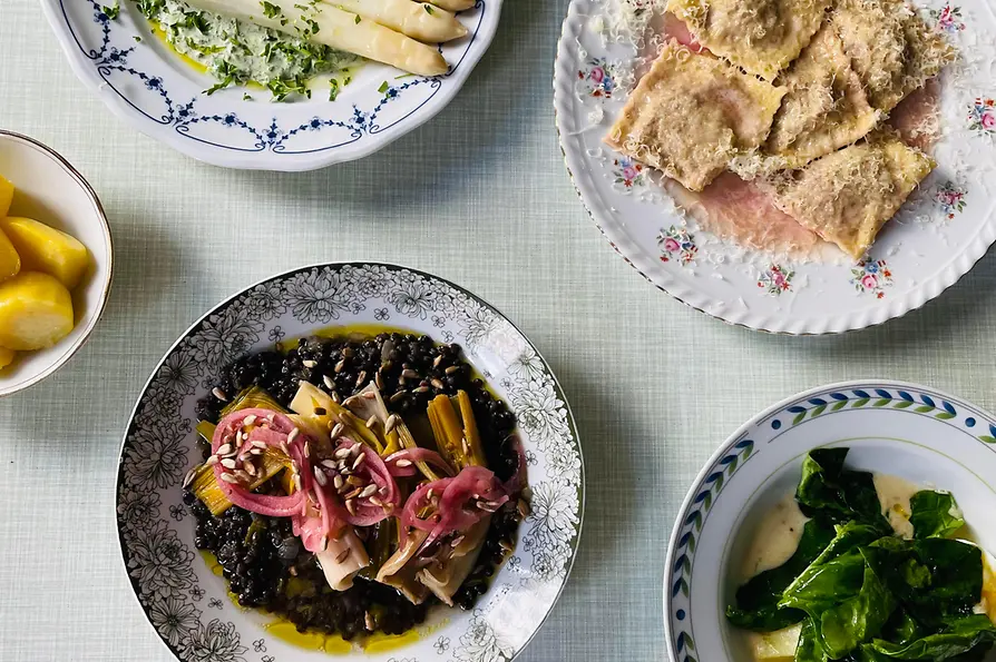 Mehrere Teller mit saisonalen Gerichten stehen auf einem hell gedeckten Tisch, darunter weißer Spargel, gefüllte Pasta und grünes Blattgemüse.