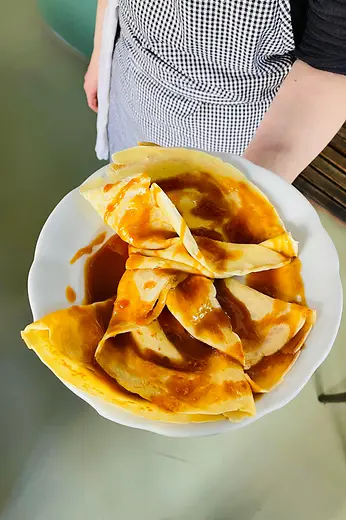 A person holds a large plate of folded crêpes generously covered in glossy caramel sauce.