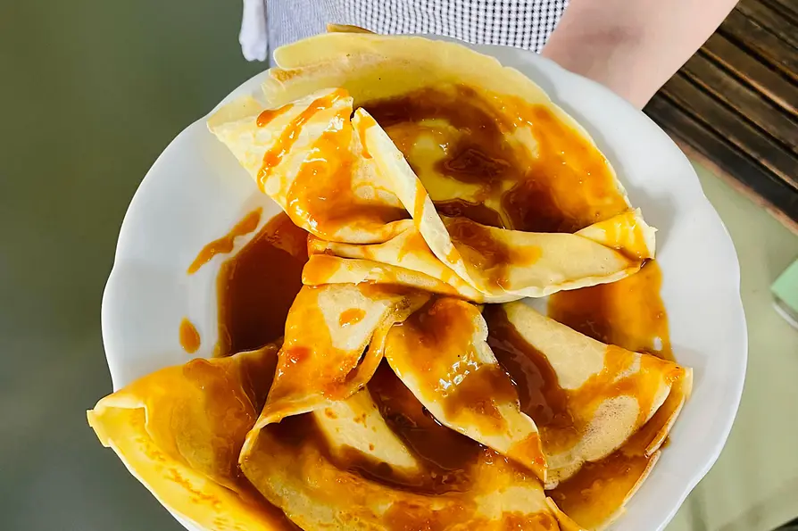 Eine Person hält einen großen Teller mit gefalteten Crêpes, die großzügig mit glänzender Karamellsauce bedeckt sind.