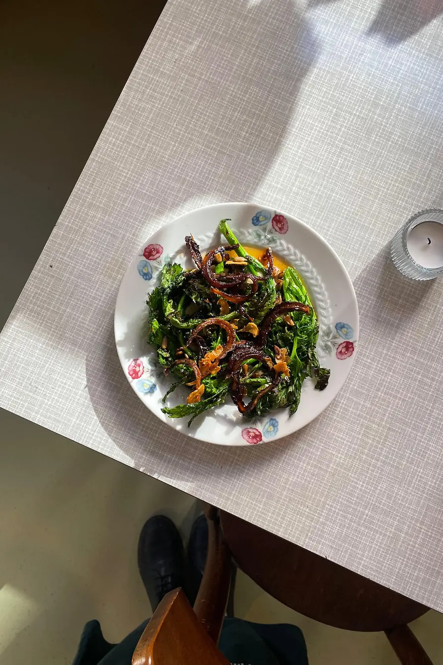 A plate of sautéed cime di rapa with caramelized onions and seeds sits in sunlight on a pale table.