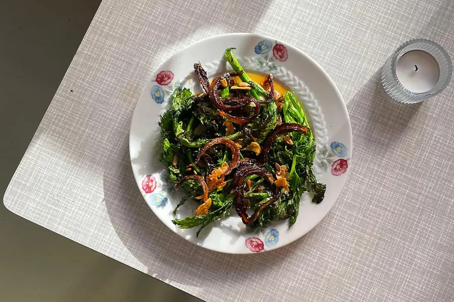 Ein Teller mit gebratenem Cime di Rapa, karamellisierten Zwiebeln und Kernen steht im Sonnenlicht auf einem hellen Tisch.