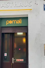Der Eingang von Pomali mit grünem Schild, dunkler Tür und dem Hinweis „ACHTUNG STUFE“ auf der unteren Glasscheibe.