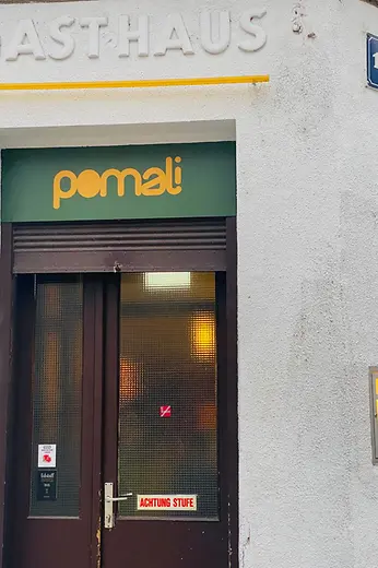The entrance to Pomali with a green sign, dark door, and an “ACHTUNG STUFE” notice on the lower glass panel.