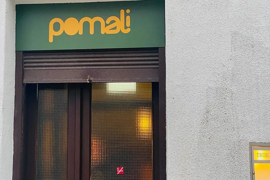 Der Eingang von Pomali mit grünem Schild, dunkler Tür und dem Hinweis „ACHTUNG STUFE“ auf der unteren Glasscheibe.