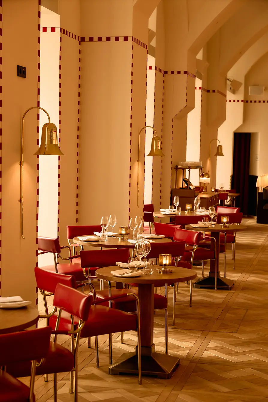Several set tables with red chairs line tall, warmly lit walls with gold-colored wall lamps.