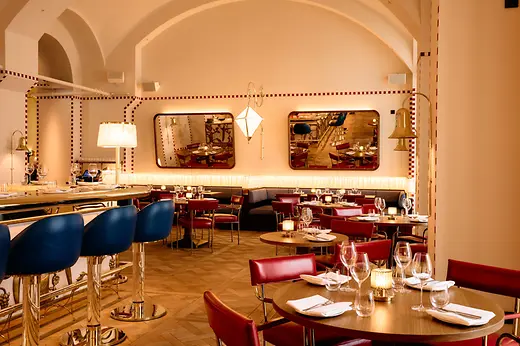 View through the dining room with round wooden tables, red chairs, wall mirrors, and soft lighting along the banquette.