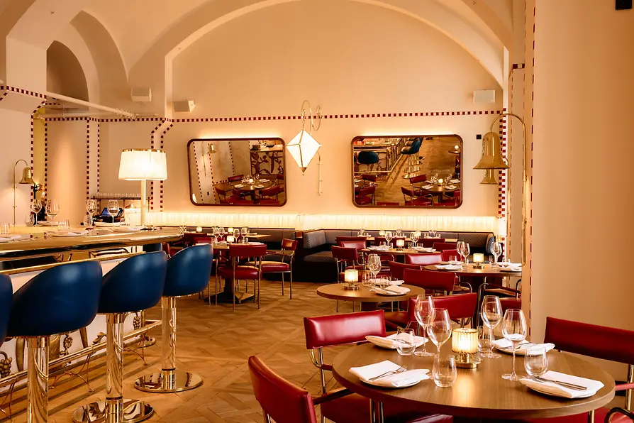 View through the dining room with round wooden tables, red chairs, wall mirrors, and soft lighting along the banquette.
