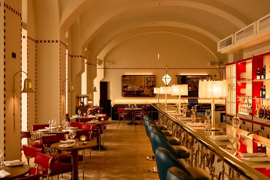 A stylish dining room with a vaulted ceiling, red chairs, blue bar stools, and a warmly lit bar.