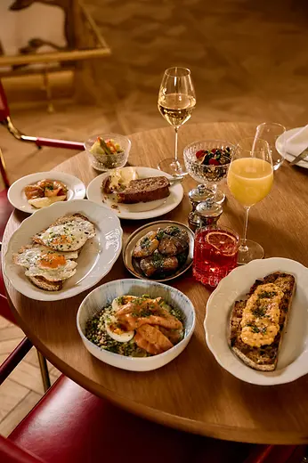 A round table is filled with brunch dishes including eggs on toast, potatoes, salmon, bread, juice, wine, and a red drink.
