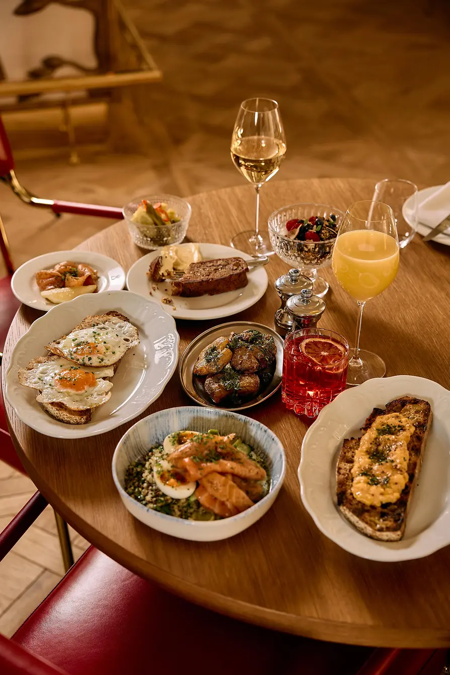 A round table is filled with brunch dishes including eggs on toast, potatoes, salmon, bread, juice, wine, and a red drink.