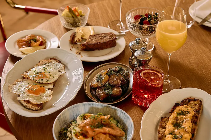 Ein runder Tisch ist mit Brunchgerichten, darunter Eier auf Toast, Kartoffeln, Lachs, Brot, Saft, Wein und ein roter Drink, reich gedeckt.