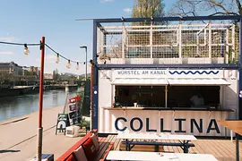Ein kleiner Kiosk mit der Aufschrift „Würstel am Kanal“ steht direkt am Wasser, davor Bänke und Lichterketten in der Sonne.