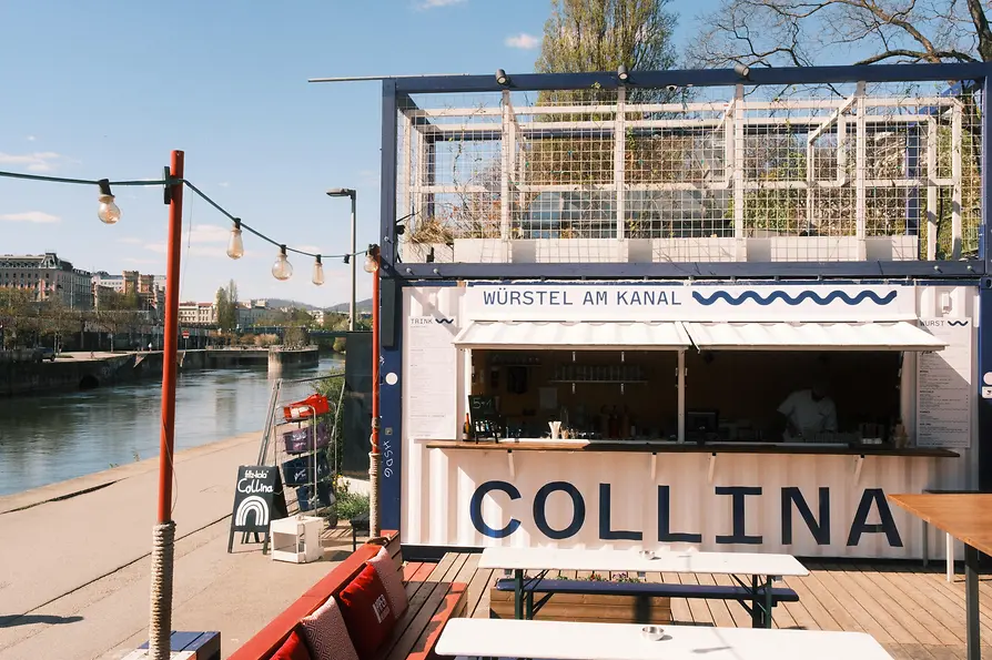 Ein kleiner Kiosk mit der Aufschrift „Würstel am Kanal“ steht direkt am Wasser, davor Bänke und Lichterketten in der Sonne.