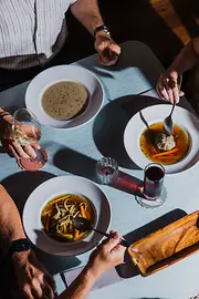 Von oben sieht man mehrere Personen an einem Tisch mit traditionellen Suppen, Wein und einem kleinen roten Getränk im Sonnenlicht.