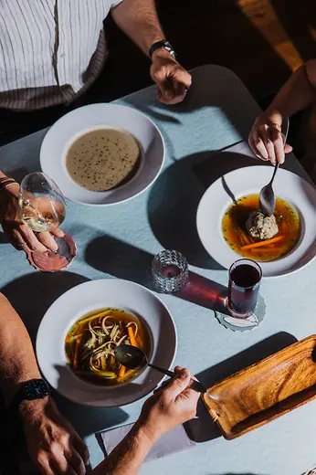 From above, several people sit at a table with traditional soups, wine, and a small red drink in the sunlight.
