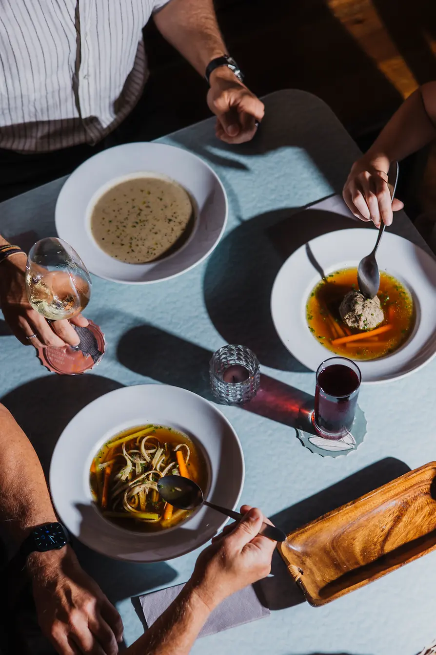 From above, several people sit at a table with traditional soups, wine, and a small red drink in the sunlight.