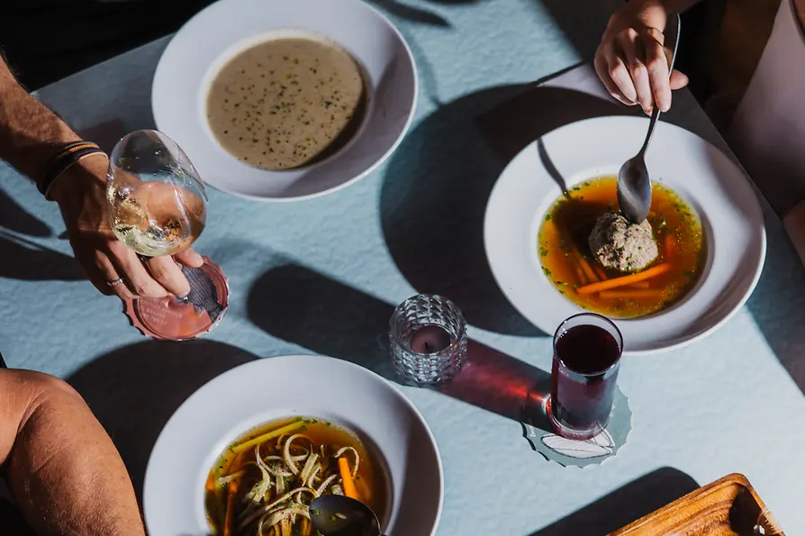 Von oben sieht man mehrere Personen an einem Tisch mit traditionellen Suppen, Wein und einem kleinen roten Getränk im Sonnenlicht.
