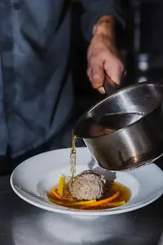 Aus einem kleinen Topf wird klare Suppe über einen Leberknödel mit Gemüsestreifen in einen tiefen Teller gegossen.