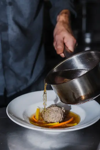 Clear broth is poured from a small pot over a liver dumpling with vegetable strips into a deep plate.