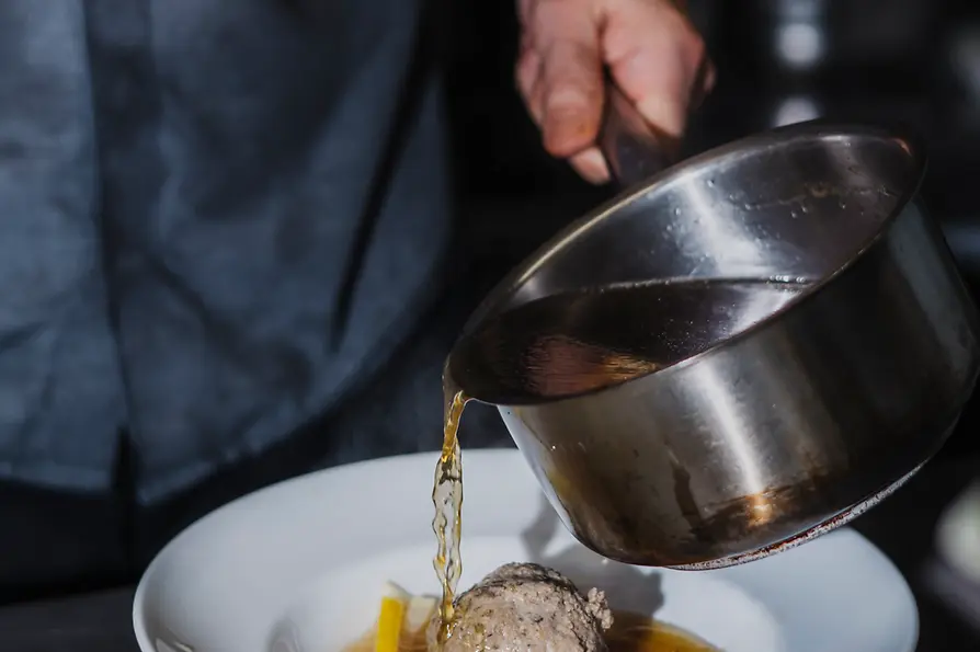 Aus einem kleinen Topf wird klare Suppe über einen Leberknödel mit Gemüsestreifen in einen tiefen Teller gegossen.