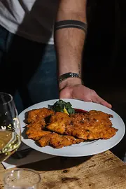 Eine Person serviert einen großen Teller mit knusprig paniertem Schnitzel auf einem Holztisch; im Vordergrund steht ein Glas Weißwein.