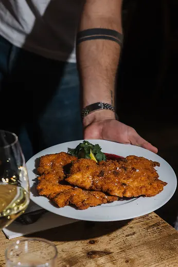 A person serves a large plate of crispy breaded schnitzel on a wooden table, with a glass of white wine in the foreground.