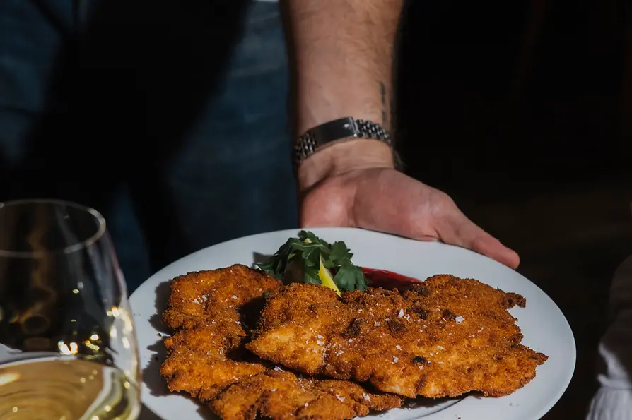 Eine Person serviert einen großen Teller mit knusprig paniertem Schnitzel auf einem Holztisch; im Vordergrund steht ein Glas Weißwein.
