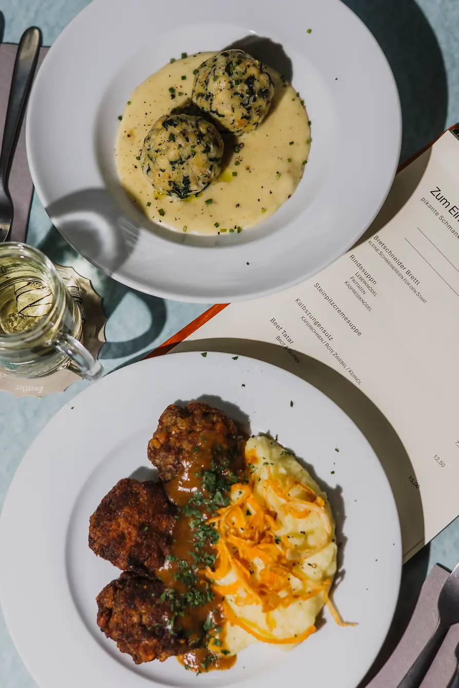 Two plates of classic dishes sit beside a menu: spinach dumplings in a light sauce and breaded meat with mashed potatoes and gravy.
