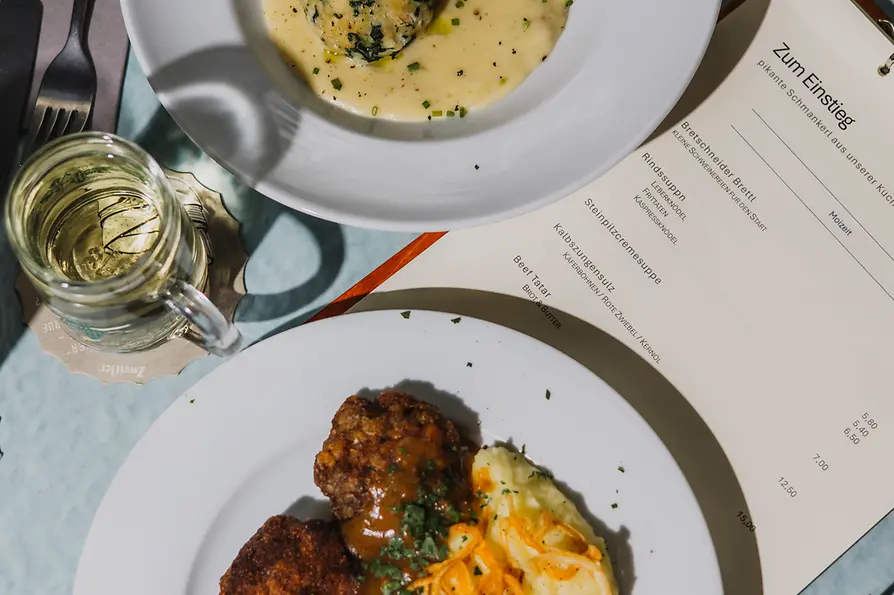 Zwei Teller mit klassischen Gerichten stehen neben einer Speisekarte: Spinatknödel in heller Sauce und paniertes Fleisch mit Püree und Sauce.