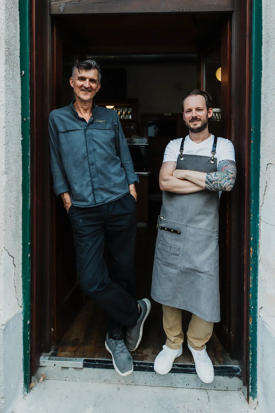 Two men stand relaxed in the restaurant doorway facing the camera; one is wearing a gray apron.