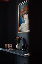 Dunkle Zimmerwand mit gerahmtem Porträt, Vase mit Blumen, Wasserkocher und Informationsmaterial auf einer Konsole.