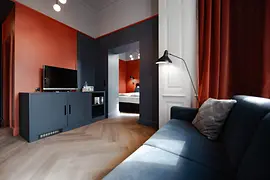 Wohnbereich einer Suite mit blauem Sofa, TV-Möbel und kräftig roten sowie dunkelblauen Wänden; im Hintergrund ist das Schlafzimmer zu sehen.