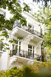 Historische helle Hausfassade mit mehreren schmiedeeisernen Balkonen, eingerahmt von grünen Bäumen und Sträuchern.