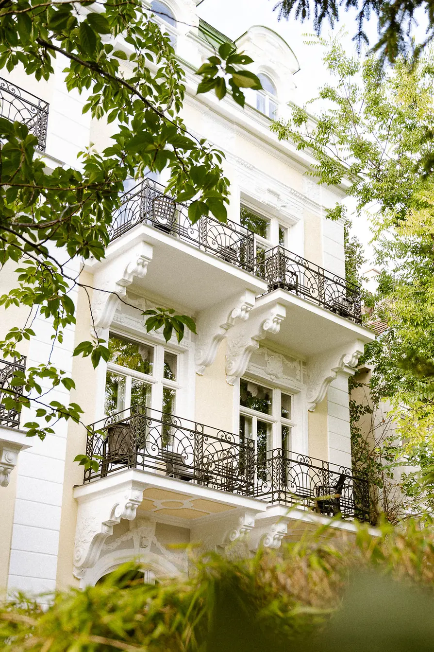 Historic light-colored facade with several wrought-iron balconies, framed by green trees and shrubs.