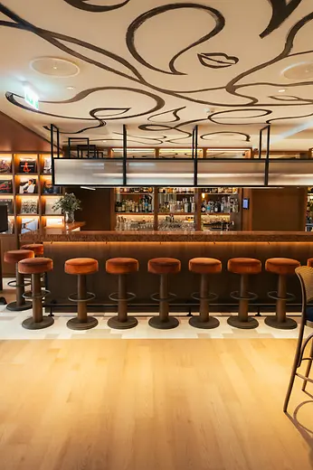 Elegant bar area with a long wooden counter, woven chairs, small table lamps, and a decorative illustrated ceiling.