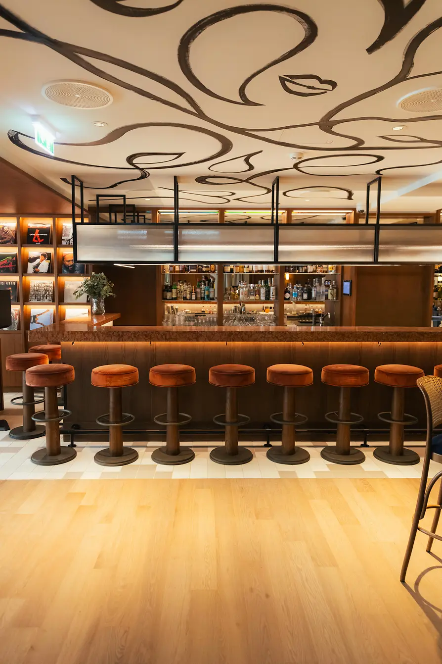 Elegant bar area with a long wooden counter, woven chairs, small table lamps, and a decorative illustrated ceiling.