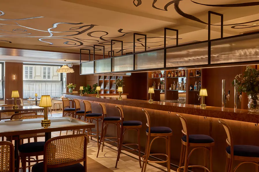 Front view of a stylish hotel bar with stools, a backlit shelf wall, and a striking graphic drawing across the ceiling.