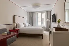 Helles Hotelzimmer mit zwei Einzelbetten, roten Beistelltischen, großem Fenster und schlichtem, elegantem Interieur.