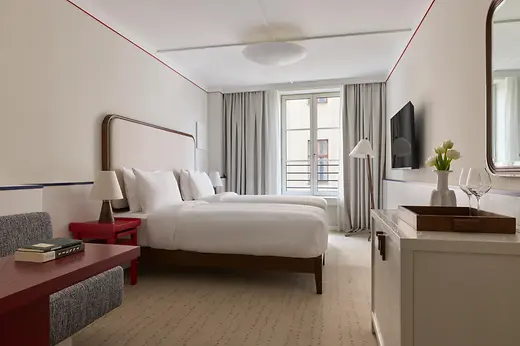 Bright hotel room with two twin beds, red side tables, a large window, and simple elegant interiors.