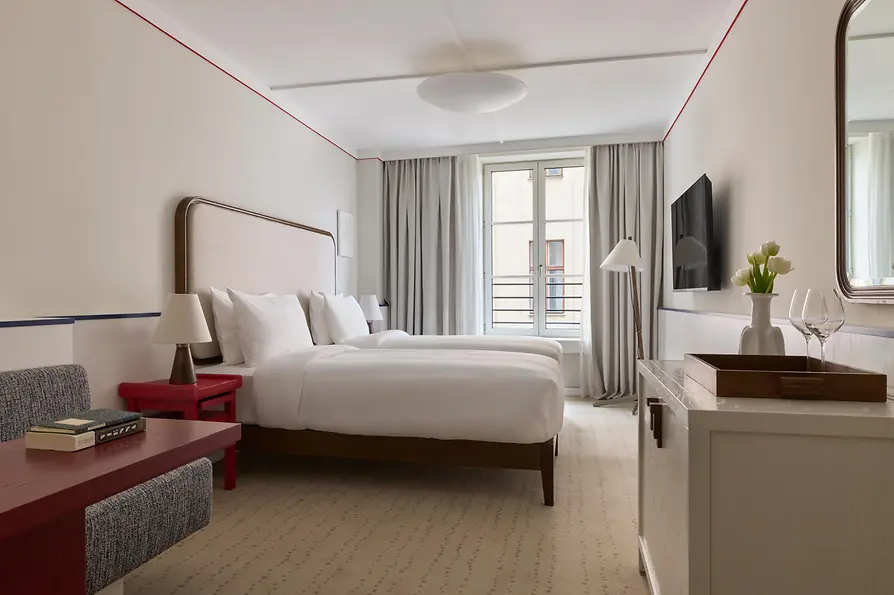 Helles Hotelzimmer mit zwei Einzelbetten, roten Beistelltischen, großem Fenster und schlichtem, elegantem Interieur.